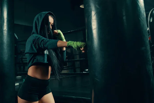 Entrenamiento de mujer atlética joven con saco de boxeo con sudadera con capucha y vendajes de lucha —  Fotos de Stock