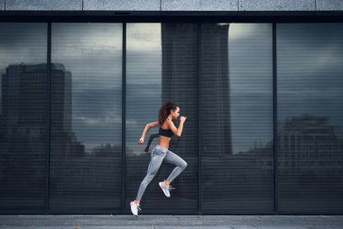 Şehir merkezinde koşarken atlayan genç atletik kadın arka planda büyük ayna pencereleri ile