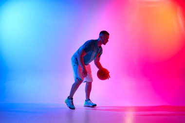 Genç atletik adam basketbol topuyla salya akıtıyor mavi ve pembe arka planda ışık yansıması ile poz veriyor.
