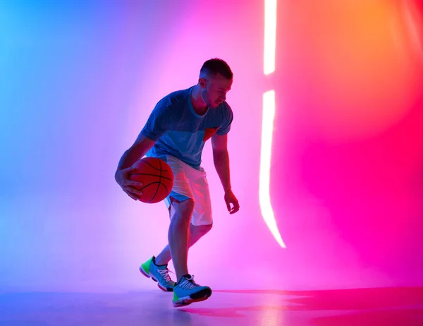 Young athletic man dribbling with basketball ball posing on mix of blue and pink background with light projection — Stock Photo, Image