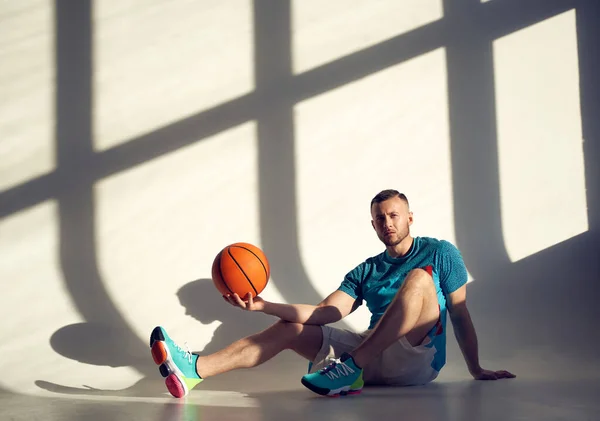 Young athletic man, basketball player holding ball with one hand and sitting near wall with shadows from window
