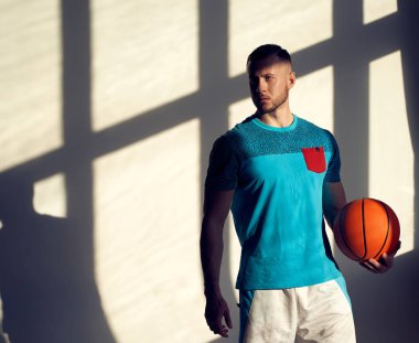 Genç atletik adam, basketbol oyuncusu topu tutuyor ve penceresinden gölgeler çıkan duvarın yanında duruyor.