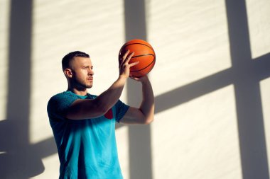 Genç atletik adam, basketbol oyuncusu topu tutuyor ve penceresinden gölgeler çıkan duvarın yanında duruyor.