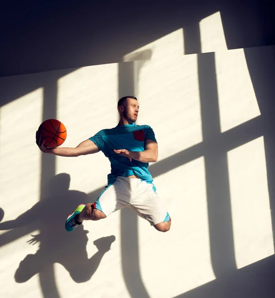 Jonge atletische basketballer springen met bal in de voorkant van de muur met schaduwen uit het raam — Stockfoto