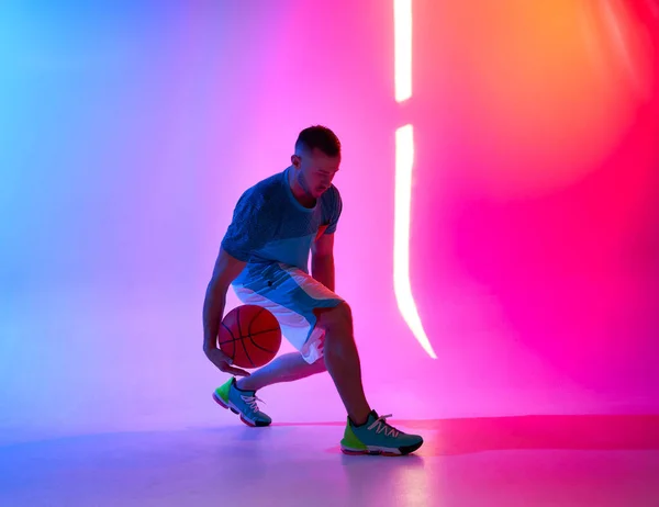 Young athletic man dribbling with basketball ball posing on mix of blue and pink background with light projection — Stock Photo, Image