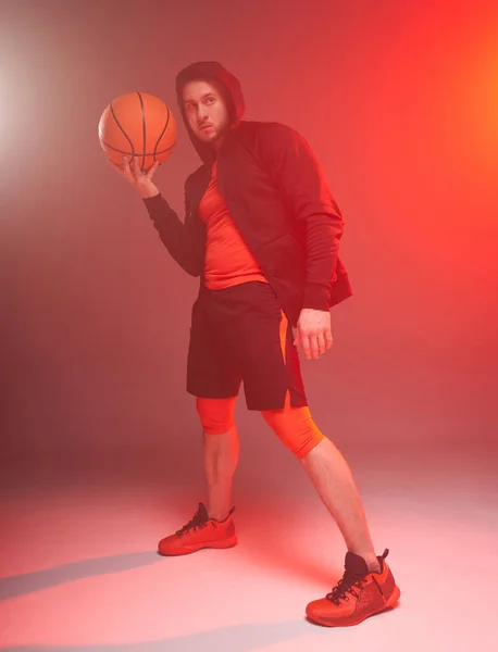 Jonge atletische man houden basketbal met een hand en het dragen van hoodie op rokerige rode achtergrond — Stockfoto