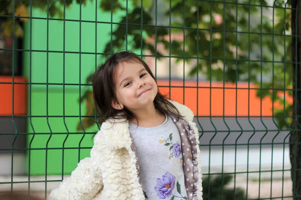 Una niña pequeña con el pelo largo y oscuro en un abrigo blanco esponjoso se para frente a una valla de metal. Fondo borroso de la calle — Foto de Stock