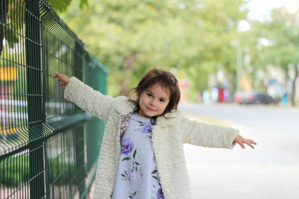 Une petite fille aux longs cheveux foncés dans un manteau blanc moelleux se tient devant une clôture métallique. Fond flou de la rue arrière — Photo