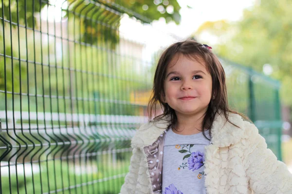 Une petite fille aux longs cheveux foncés dans un manteau blanc moelleux se tient devant une clôture métallique. Fond flou de la rue arrière — Photo