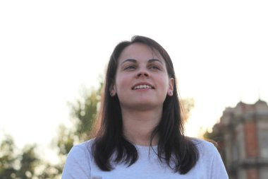 Gülümseyen, uzun saçlı, normal tişörtlü beyaz kız, akşam güneşinin arka planındaki ışıklara bakıyor.