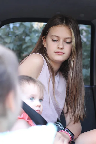 ピンクのTシャツを着た小さな笑顔の子供は 車のシートベルトで固定されて座っています 背景に大人の長髪の妹は女の子を締結します — ストック写真