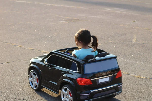 Küçük kuyruklu kız şehirde bir meydanda asfalt üzerinde siyah bir oyuncak araba kullanıyor.