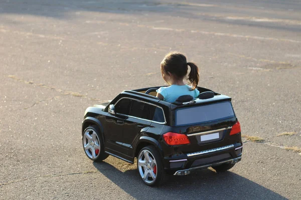 Küçük kuyruklu kız şehirde bir meydanda asfalt üzerinde siyah bir oyuncak araba kullanıyor.
