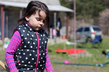 Beyaz saçlı, açık havada at kuyruklu beyaz bir kız ellerinde uzun tahta bir sopa tutuyor. Anaokulu öğrencisi bahar bahçesinde ebeveynlere yardım ediyor.