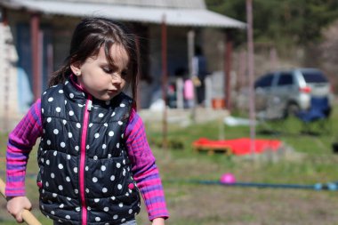 Beyaz saçlı, açık havada at kuyruklu beyaz bir kız ellerinde uzun tahta bir sopa tutuyor. Anaokulu öğrencisi bahar bahçesinde ebeveynlere yardım ediyor.