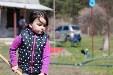 Beyaz saçlı, açık havada at kuyruklu beyaz bir kız ellerinde uzun tahta bir sopa tutuyor. Anaokulu öğrencisi bahar bahçesinde ebeveynlere yardım ediyor.