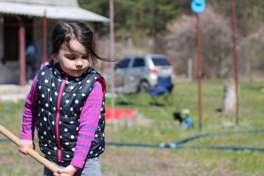 Beyaz saçlı, açık havada at kuyruklu beyaz bir kız ellerinde uzun tahta bir sopa tutuyor. Anaokulu öğrencisi bahar bahçesinde ebeveynlere yardım ediyor.