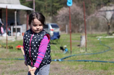 Beyaz saçlı, açık havada at kuyruklu beyaz bir kız ellerinde uzun tahta bir sopa tutuyor. Anaokulu öğrencisi bahar bahçesinde ebeveynlere yardım ediyor.
