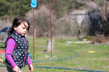 Beyaz saçlı, açık havada at kuyruklu beyaz bir kız ellerinde uzun tahta bir sopa tutuyor. Anaokulu öğrencisi bahar bahçesinde ebeveynlere yardım ediyor.
