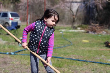 Beyaz saçlı, açık havada at kuyruklu beyaz bir kız ellerinde uzun tahta bir sopa tutuyor. Anaokulu öğrencisi bahar bahçesinde ebeveynlere yardım ediyor.