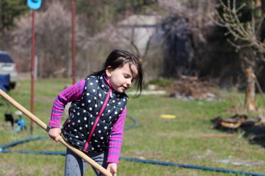 Beyaz saçlı, açık havada at kuyruklu beyaz bir kız ellerinde uzun tahta bir sopa tutuyor. Anaokulu öğrencisi bahar bahçesinde ebeveynlere yardım ediyor.