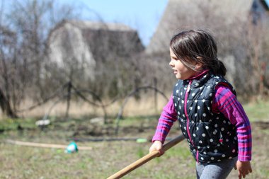 Beyaz saçlı, açık havada at kuyruklu beyaz bir kız ellerinde uzun tahta bir sopa tutuyor. Anaokulu öğrencisi bahar bahçesinde ebeveynlere yardım ediyor.