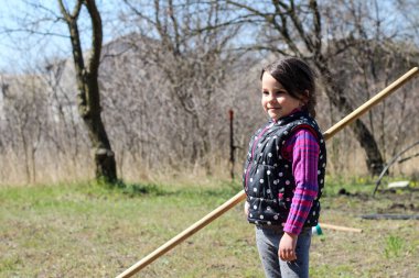 Beyaz saçlı, açık havada at kuyruklu beyaz bir kız ellerinde uzun tahta bir sopa tutuyor. Anaokulu öğrencisi bahar bahçesinde ebeveynlere yardım ediyor.