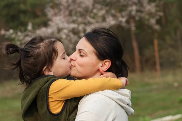 美しい母親と娘はカジュアルな服で抱き合ってキスしています ぼんやりとした開花木の背景に暖かい赤ちゃん抱擁 — ストック写真
