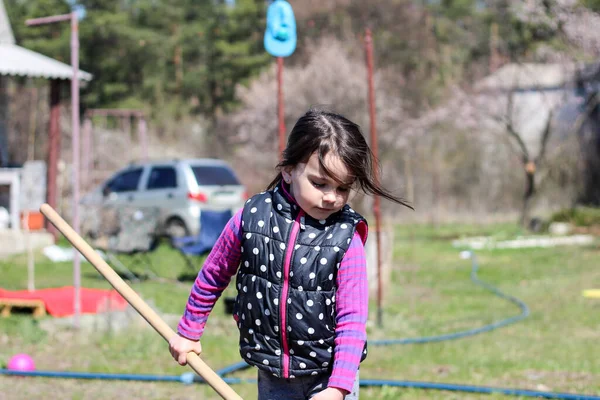 Beyaz saçlı, açık havada at kuyruklu beyaz bir kız ellerinde uzun tahta bir sopa tutuyor. Anaokulu öğrencisi bahar bahçesinde ebeveynlere yardım ediyor.