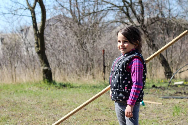 Beyaz saçlı, açık havada at kuyruklu beyaz bir kız ellerinde uzun tahta bir sopa tutuyor. Anaokulu öğrencisi bahar bahçesinde ebeveynlere yardım ediyor.