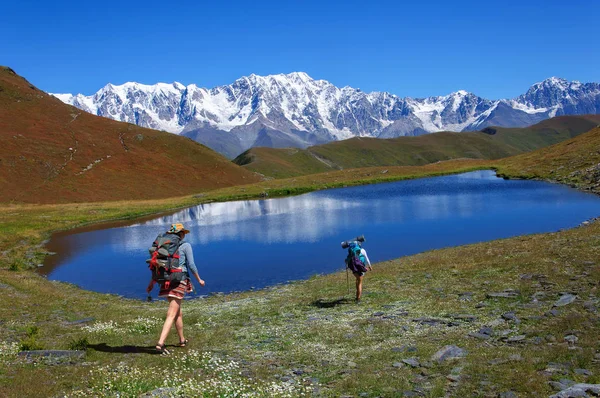 Girls with large backpacks descend to picturesque lake in the mountains of Georgia — Stock Photo, Image