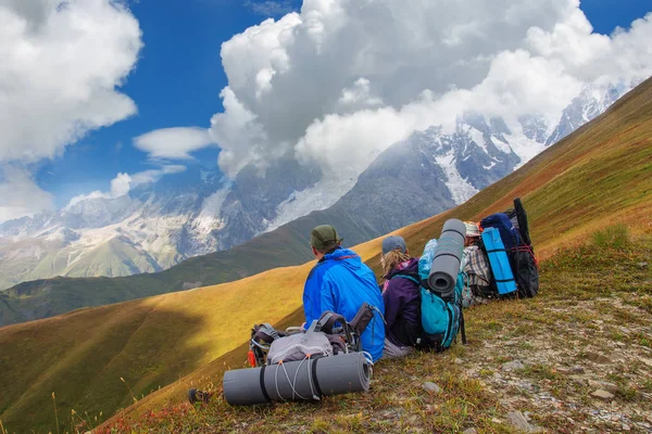 Група туристів з великими рюкзаками відпочиває на зеленому лузі з видом на гори — стокове фото