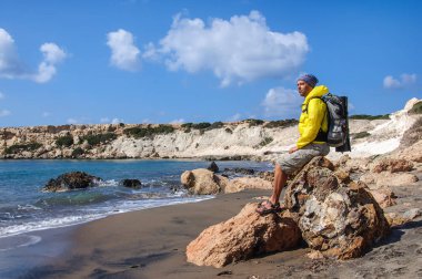 Sırt çantalı bir adam kayanın üzerinde oturuyor ve Akdeniz 'e bakıyor.