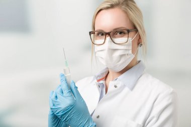 Steteskop ve tıbbi maskeli kadın doktor arka planda klinik odası olan bir şırınga tutuyor.