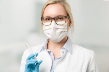 Steteskop ve tıbbi maskeli kadın doktor arka planda klinik odası olan bir şırınga tutuyor.