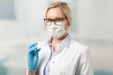 Steteskop ve tıbbi maskeli kadın doktor arka planda klinik odası olan bir şırınga tutuyor.