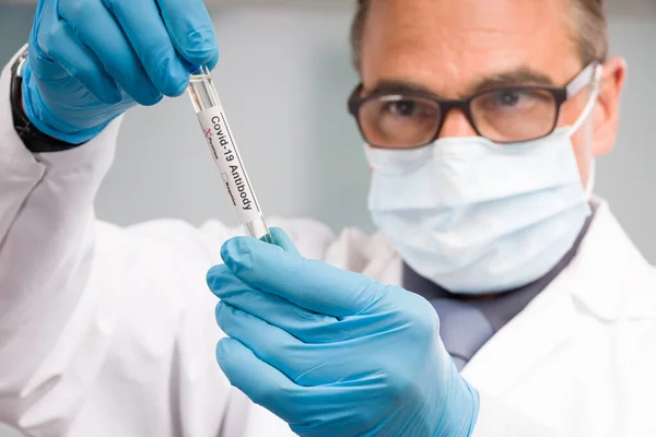 Scientist or doctor with medical face mask and medical gloves is handling virus antibody test tube with result markers in front of laboratory in background