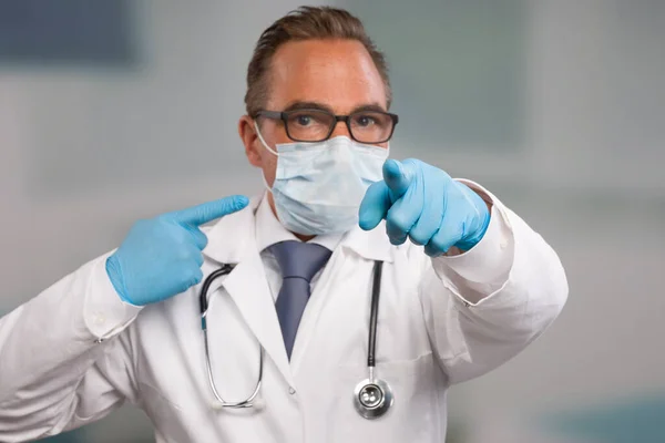 Male family doctor in doctor\'s overall and tie with stethoscope, medical gloves and medical face mask points to his face mask and directly