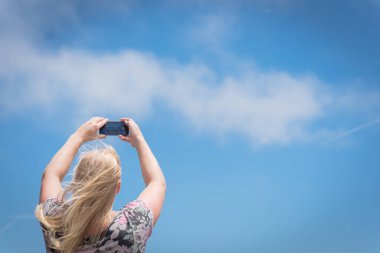 Akıllı telefonuyla fotoğraf çeken beyaz bir bayanın görüntüsüne bak. Düşük açılı sarışın kız cep telefonu ile bulutların altında mavi gökyüzü