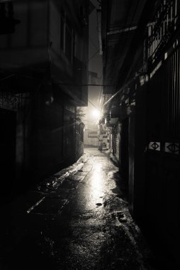 Hanoi, Vietnam 'da gece vakti karanlık, gölgeli ve tehlikeli görünen bir arka sokak. Uzun yolun sonundaki lambanın ıslak kaldırımına alçak ışık yansıyor.