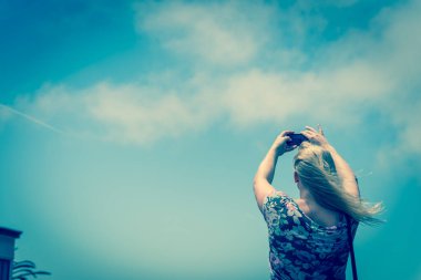 Beyaz bir bayanın akıllı telefonuyla fotoğraf çekerken çekilmiş klasik ses tonu. Düşük açılı sarışın kız omuz askısı takıyor. Cep telefonunu yaz bulutlarının altında kullan. Mavi gökyüzü.