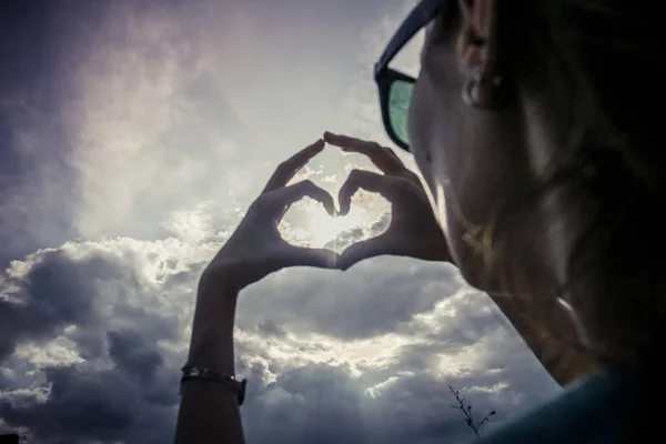 Woman making heart shape with hands against clouds