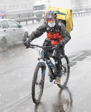 Ukrayna Kiev 23 Mart 2020: Uber 'in bir çalışanı bisikletle yiyecek dağıtıyor. Dışarısı soğuk ve kar yağıyor. Yüz, yeni COVID-19 veya SARS-CoV-2 koronavirüsünden etkilenen bölgede faaliyet gösterdiği için bir maske ile korunuyor..