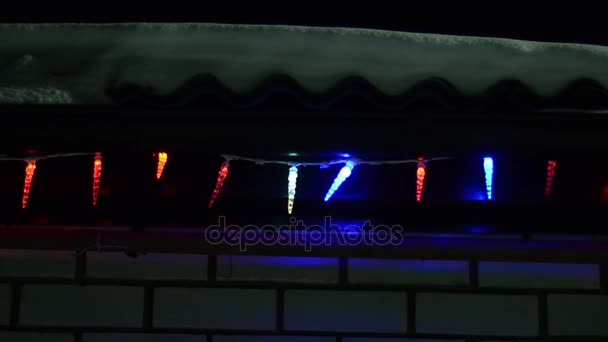 Luces de Navidad multicolores decoran la casa por la noche, guirnalda brillante en el techo de invierno, nieve yace en el techo . — Vídeos de Stock