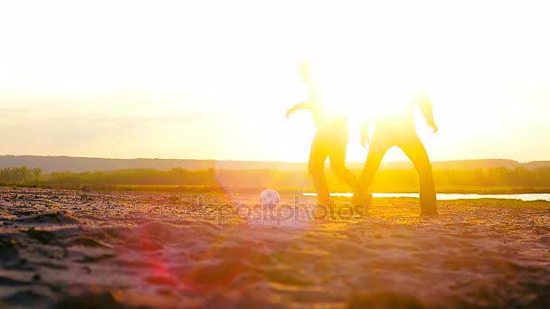 Hombres jugando al fútbol en la playa al atardecer, pelota de fútbol rodando en la arena al sol . — Vídeos de Stock