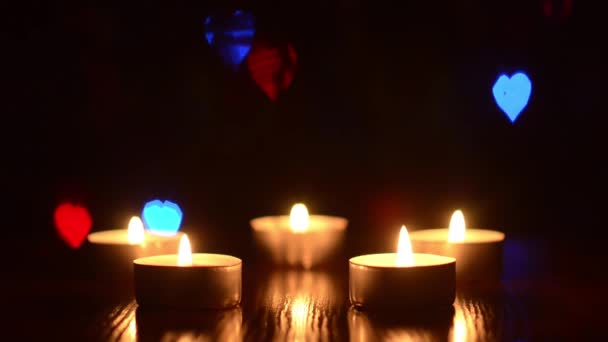 Queimando velas em uma mesa em um quarto escuro, piscando corações coloridos bokeh azul vermelho verde, decoração festiva do quarto . — Vídeo de Stock