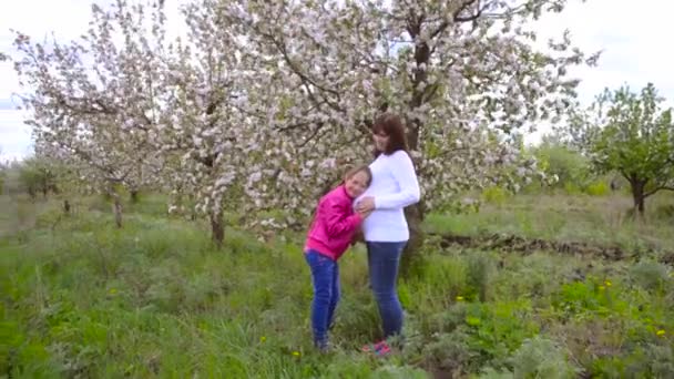 Madre embarazada abrazando a su hija en el florecido parque de primavera. Hija abraza vientre de una madre embarazada . — Vídeos de Stock