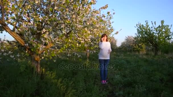 Gravid kvinne smiler i telefonen i blomstrende park om våren, jente går i blomstrende Eplehage – stockvideo