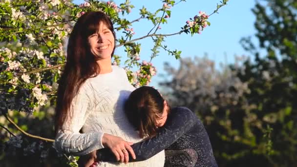 Mujer embarazada riendo, abrazando a su hija en el florecido parque de primavera. Hija abraza el vientre de la madre embarazada . — Vídeo de stock