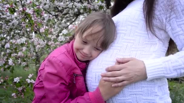 Junges Mädchen lächelnd umarmt Bauch der schwangeren Mutter. Schwangere Mutter umarmt Tochter im blühenden Frühlingspark. — Stockvideo